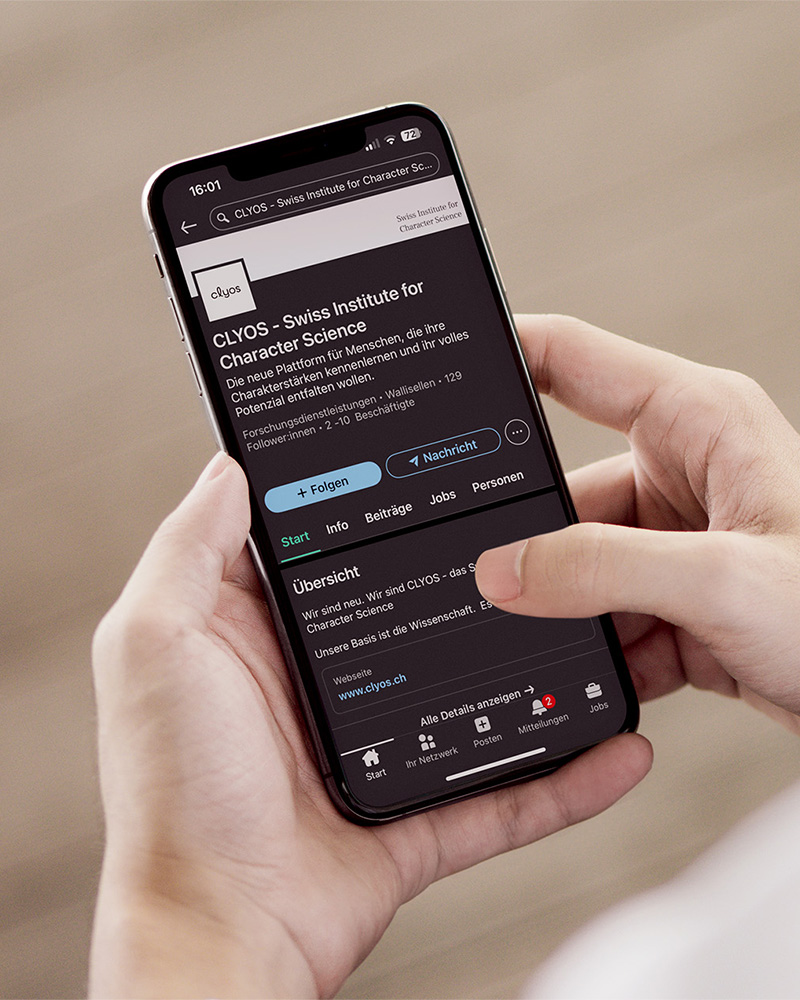 Clyos LinkedIn Person hält ein Smartphone in der Hand. Geöffnet ist die LinkedIn-Seite von Clyos – Swiss Institute for Character Science.